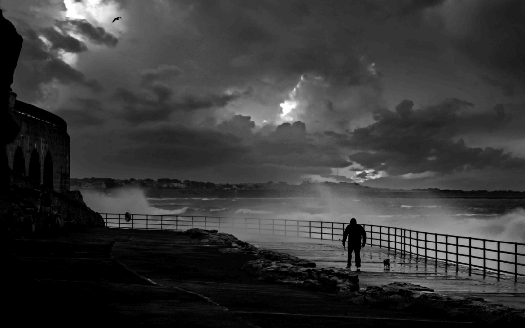 Людина з собакою йде вздовж набережної під час шторму. Великі хвилі б’ються об берег, темні хмари закривають небо. Атмосфера кризи, сили стихії та пошуку опори.